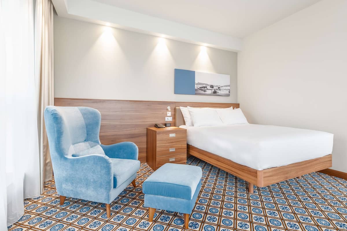 Ein Hotelzimmer mit einem Bett, einem blauen Sessel, einem Nachttisch aus Holz und einem gemusterten Boden.