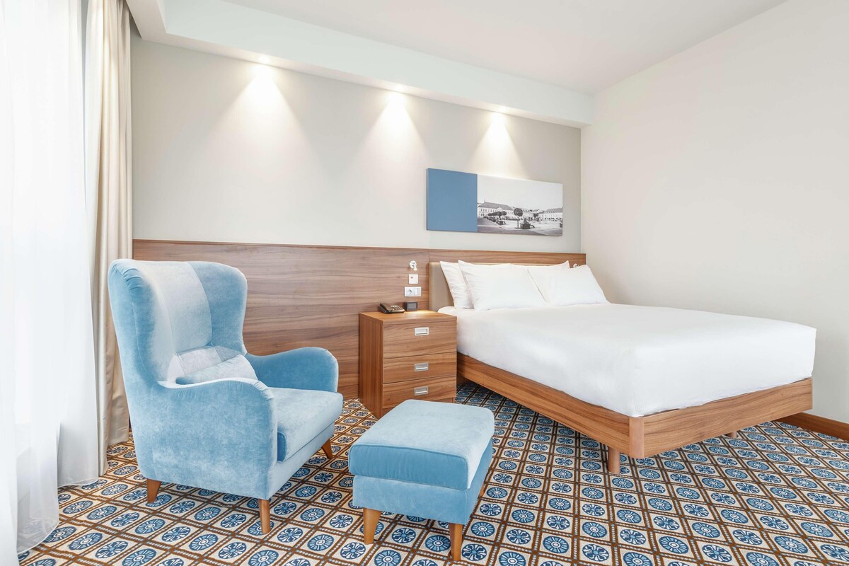 A hotel room with a bed, a nightstand, a blue armchair, and a matching ottoman.