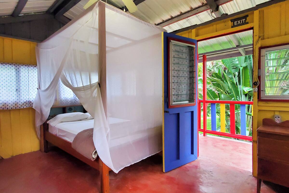A room with a four-poster bed, a small table, and a bench. There is a door with a sign that reads "EXIT" and a window with a view of greenery outside.