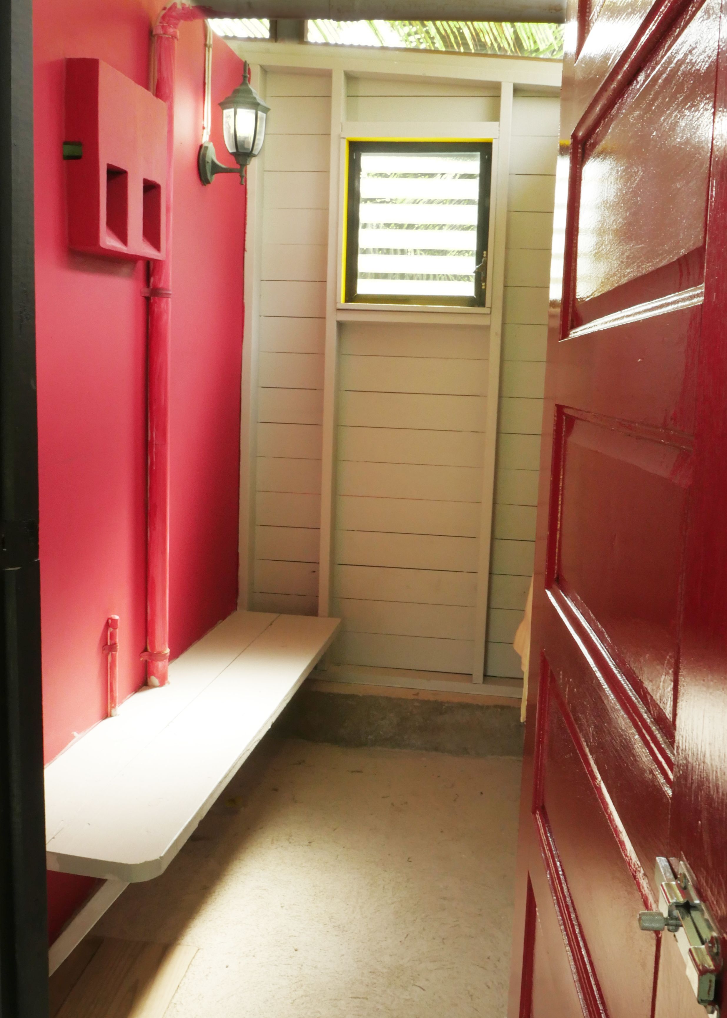 A narrow hallway with a red door on the right, a white bench on the left, and a window with horizontal blinds in the center.