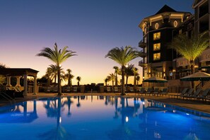 A swimming pool with lounge chairs and palm trees, surrounded by a building with multiple balconies and a covered area.