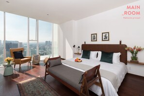 A spacious main room with a large bed, a chair, a small table with flowers, and a city view through large windows.