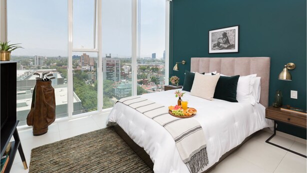 A bedroom with a large window, a bed with a tray of fruit, and a view of the city. - Monet Tower By The Local Way (Mexico City)