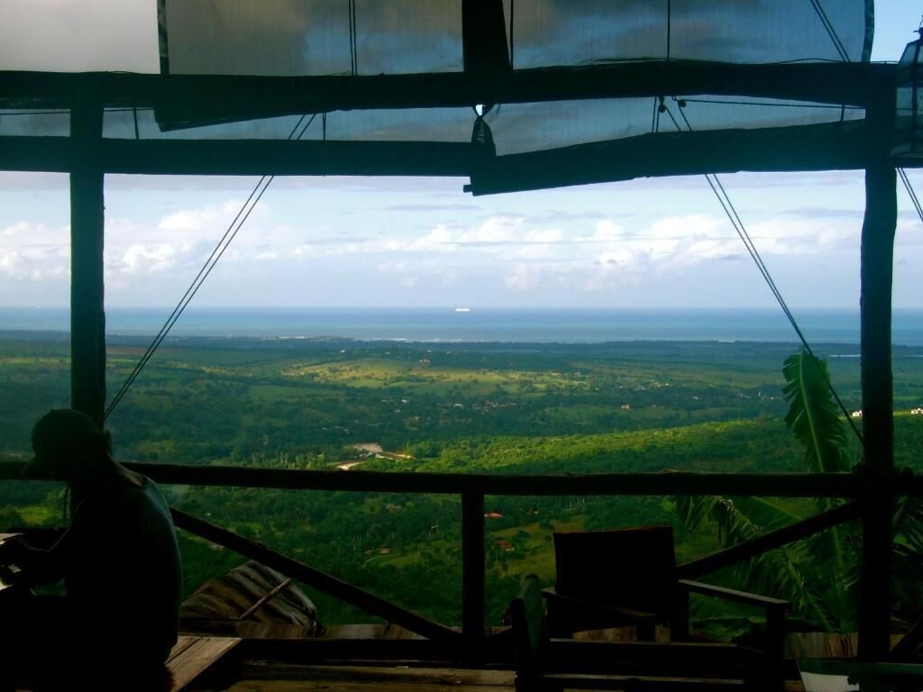A view from a covered outdoor area with a wooden railing, overlooking a green landscape with distant buildings.