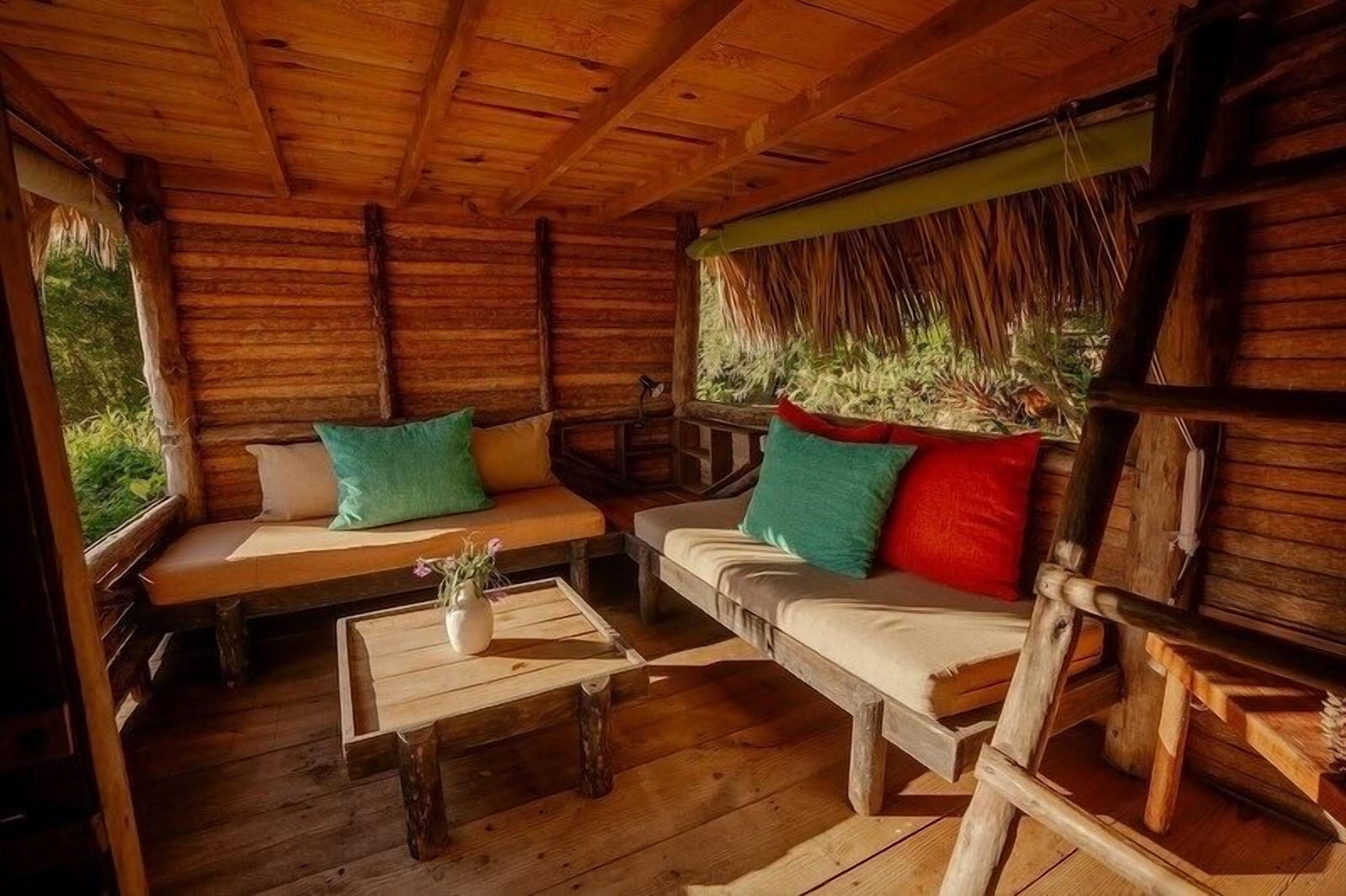 A rustic wooden cabin interior with a wooden bench, a sofa with cushions, and a small table with a vase.