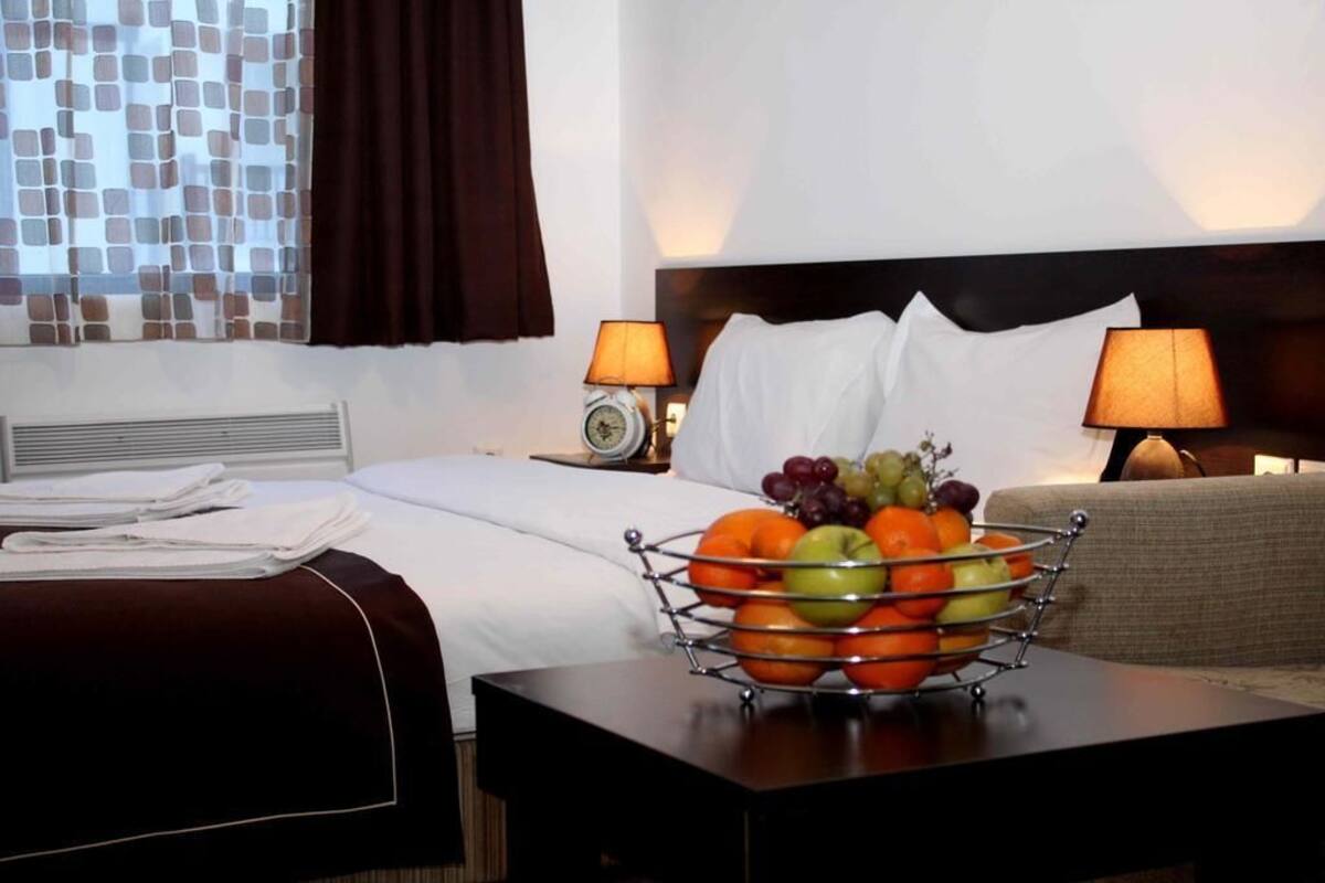 Una habitación de hotel con una cama bien hecha, una mesita de noche con frutero, dos lámparas y una ventana con cortinas.