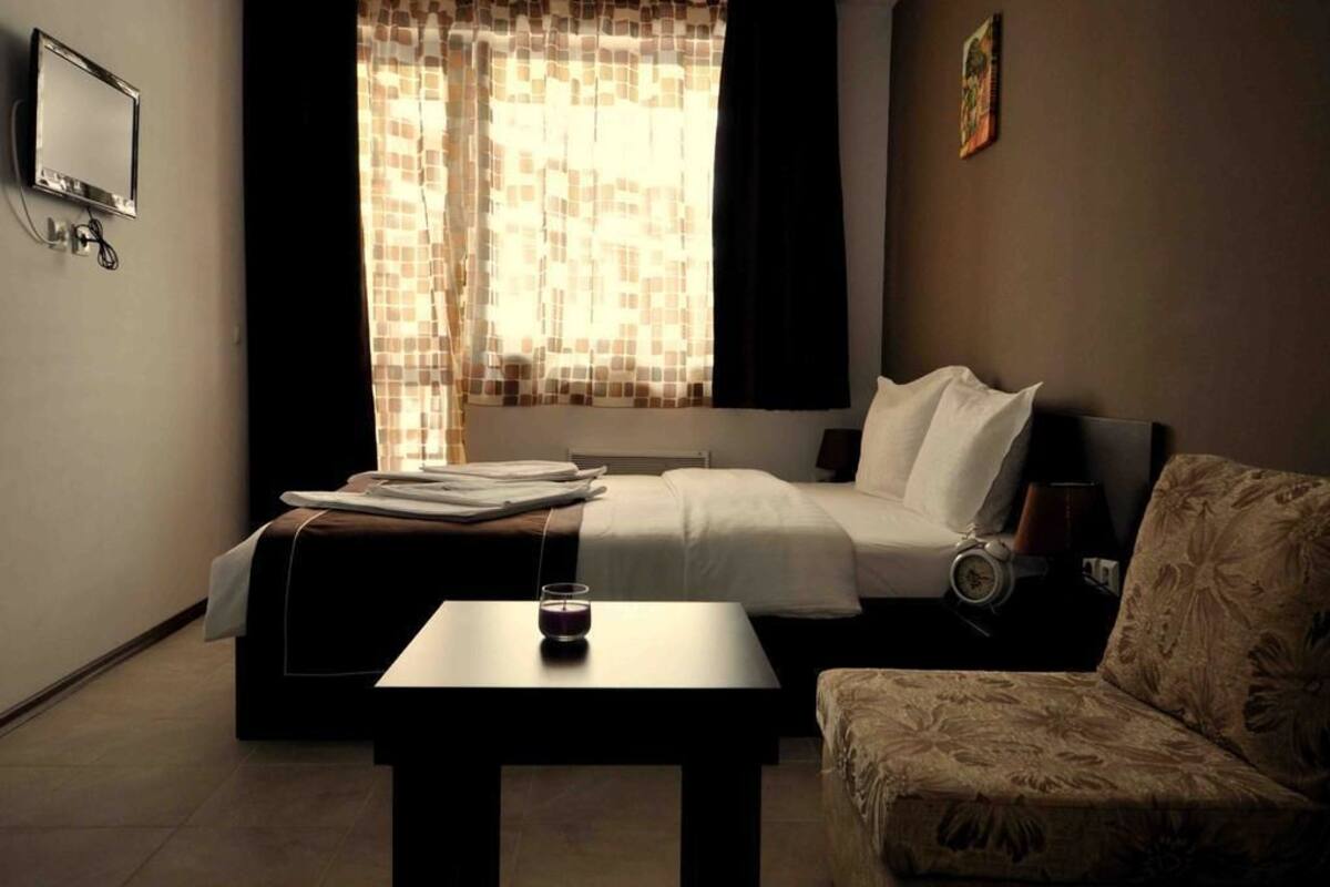 Habitación de hotel con cama, mesita de noche con lámpara, una silla, un reloj y una ventana con cortinas.