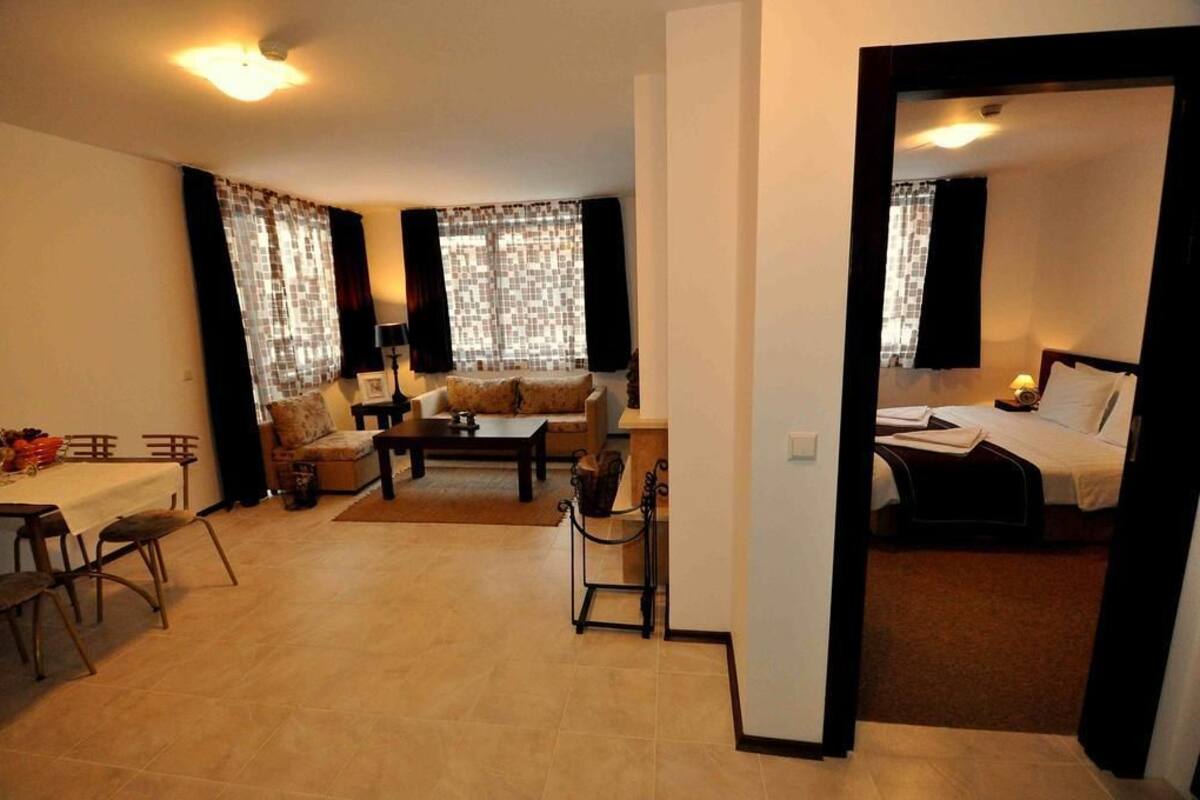 Habitación de hotel compacta con cama, una pequeña zona de estar con sofá y una mesa de centro, una mesa de comedor con sillas, y vistas a otra habitación por una puerta abierta.