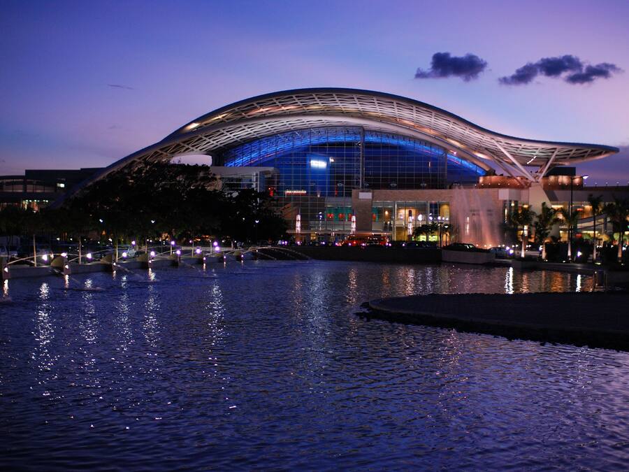 A modern architectural building with a curved roof, a reflective water body in front, and surrounding urban infrastructure.