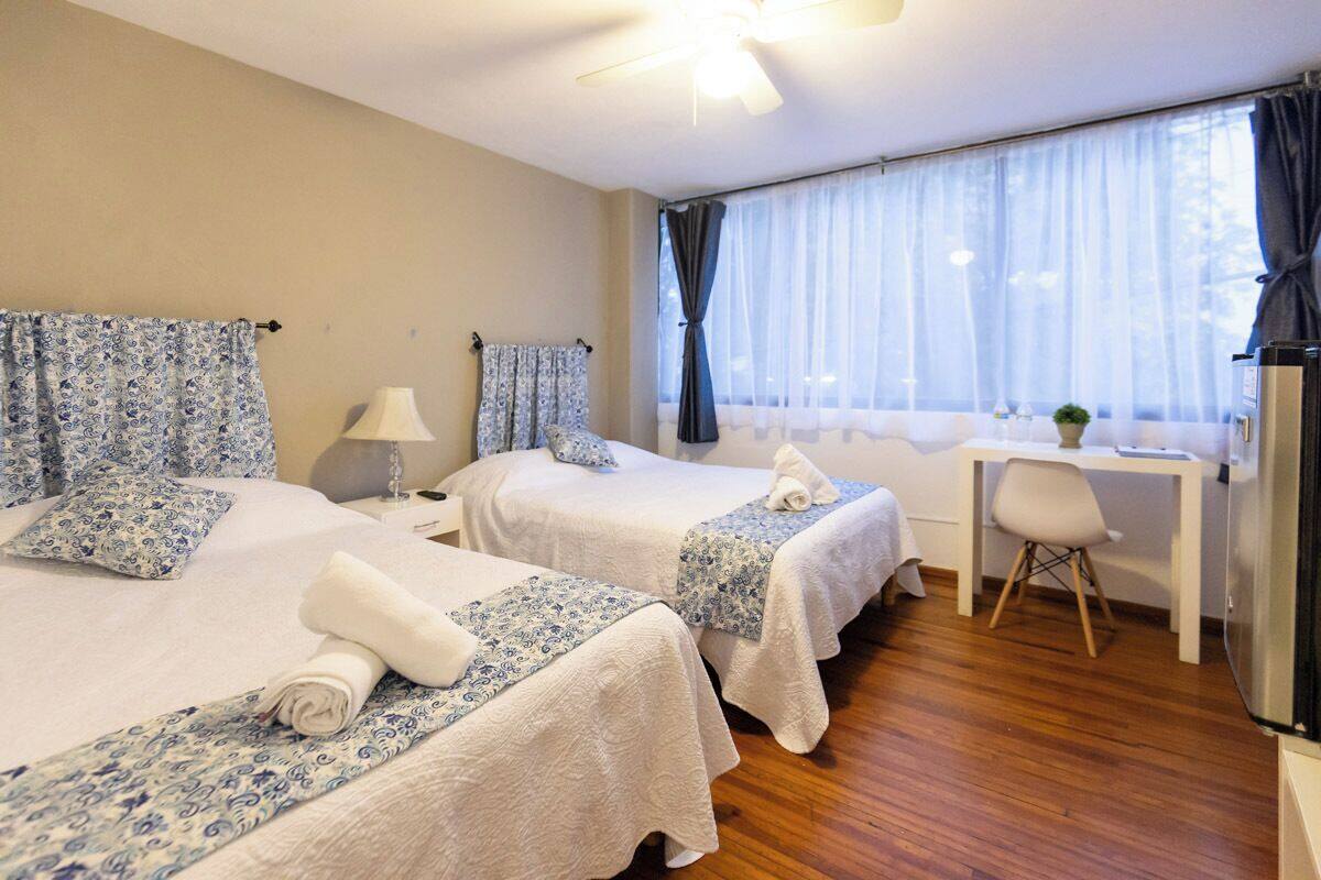 A hotel room with two beds, a desk, a chair, a ceiling fan, and a window with curtains.