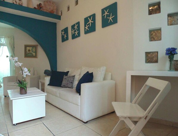 A living room with a white sofa, a white coffee table, and a white chair. There are decorative starfish on the wall and a vase with flowers on a shelf.