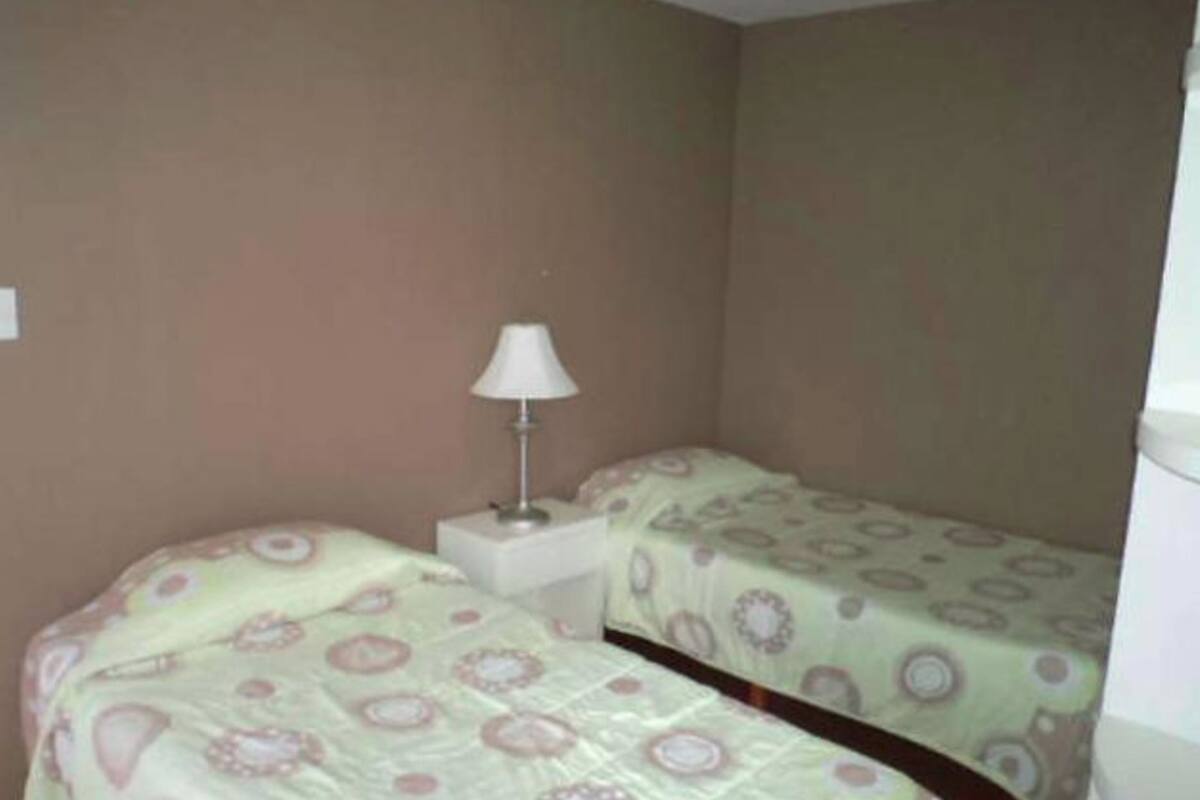 A small, single-room hotel room with two beds, a nightstand, and a lamp.