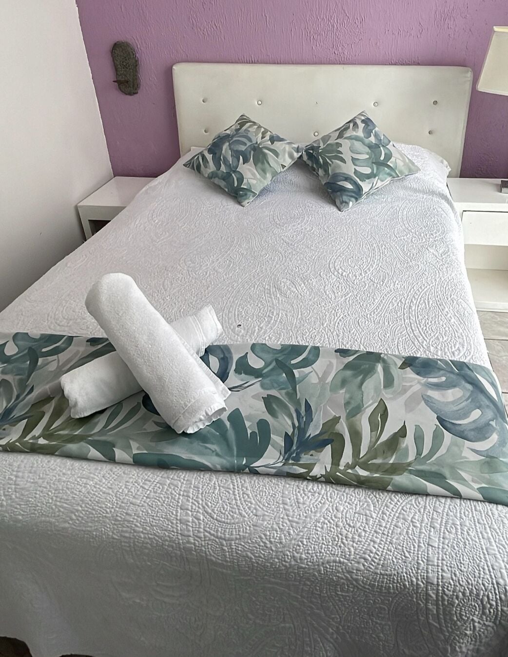 A neatly made bed with a floral-patterned bedspread, a rolled white towel, and matching pillows.