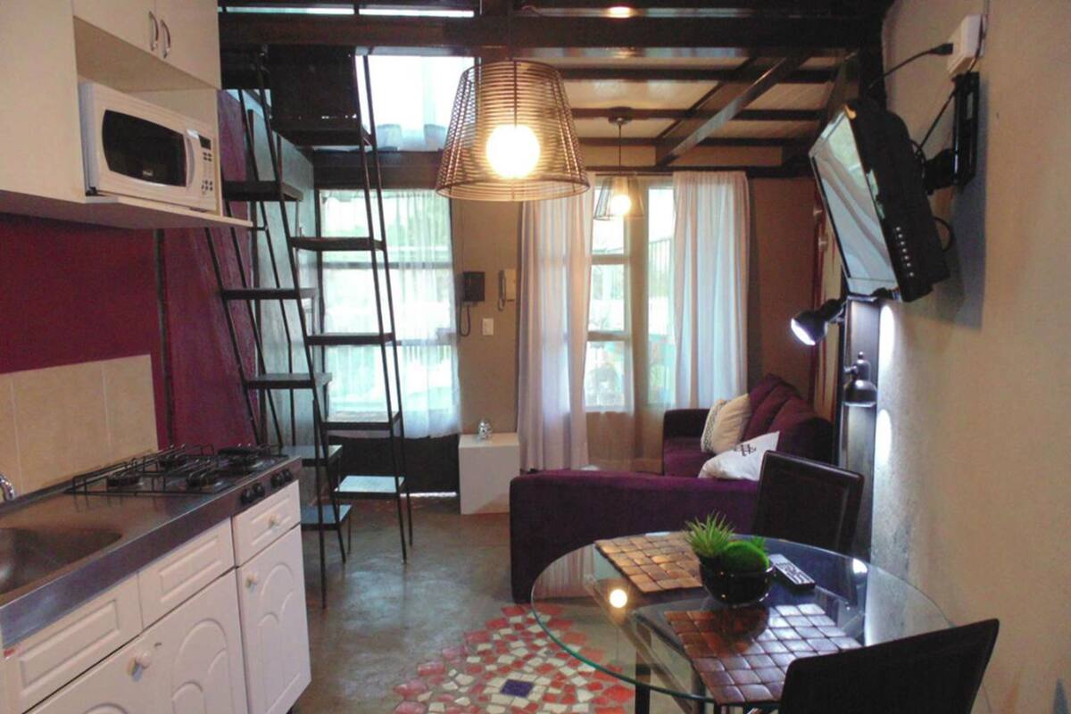 A compact kitchen with a microwave, stove, and washing machine. There is a dining area with a table and chairs, a sofa, and a small potted plant.