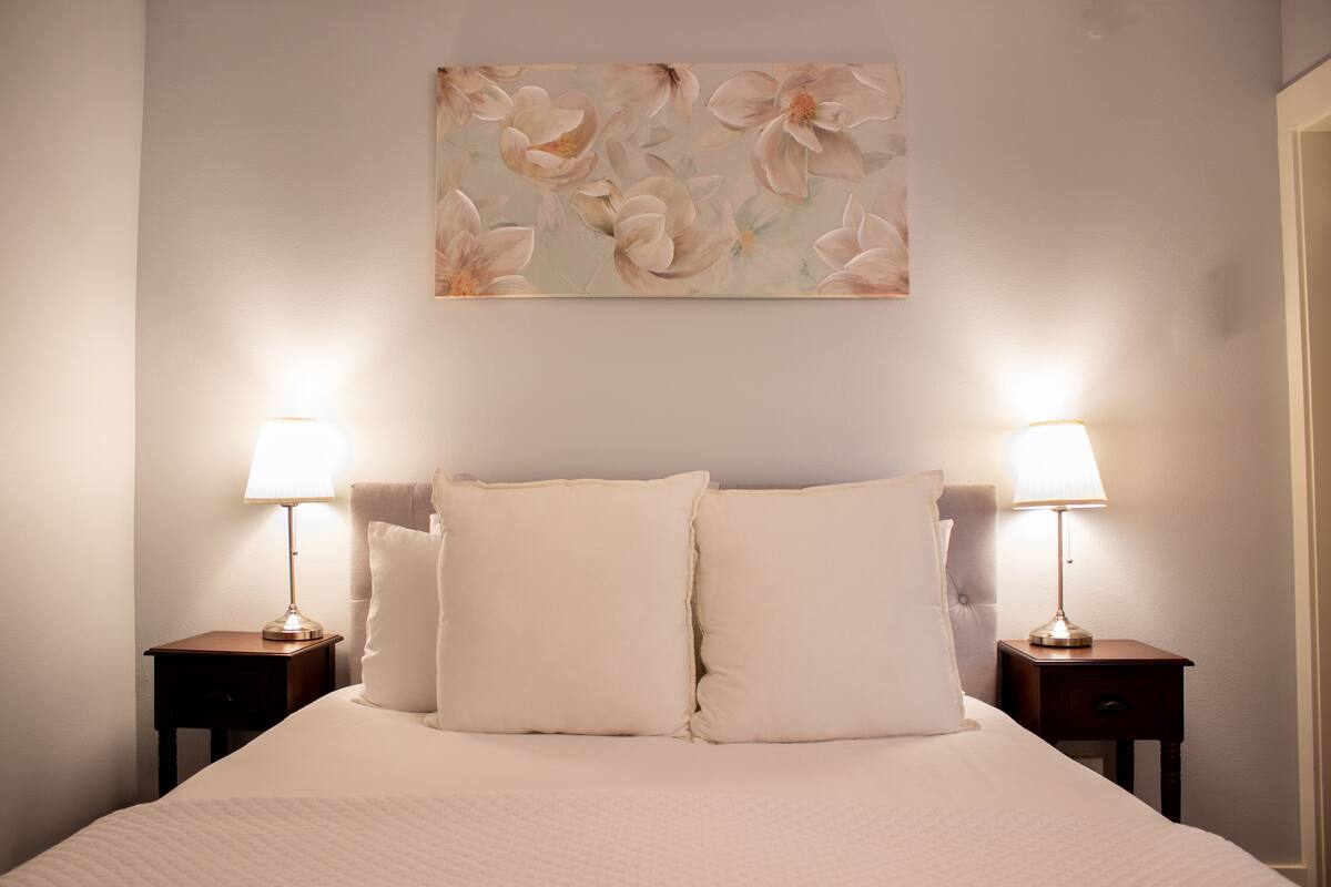 A neatly made bed with two bedside tables, each with a lamp, and a floral painting above the bed.