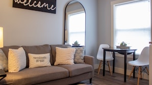 Un salon comprenant un canapé, une table basse et une table à manger avec des chaises. Un grand miroir orne le mur, et une fenêtre offre une vue dégagée.