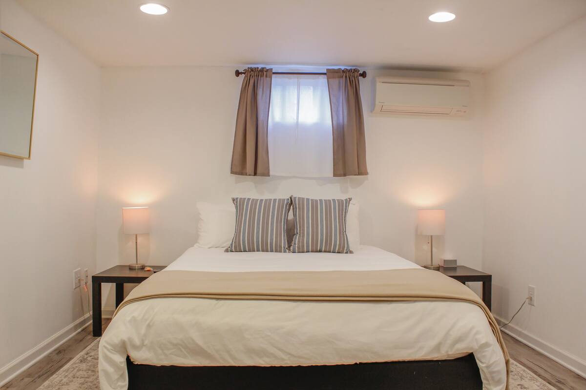 A bedroom with a bed, two bedside tables, a window with curtains, an air conditioning unit, and a wall-mounted light fixture.