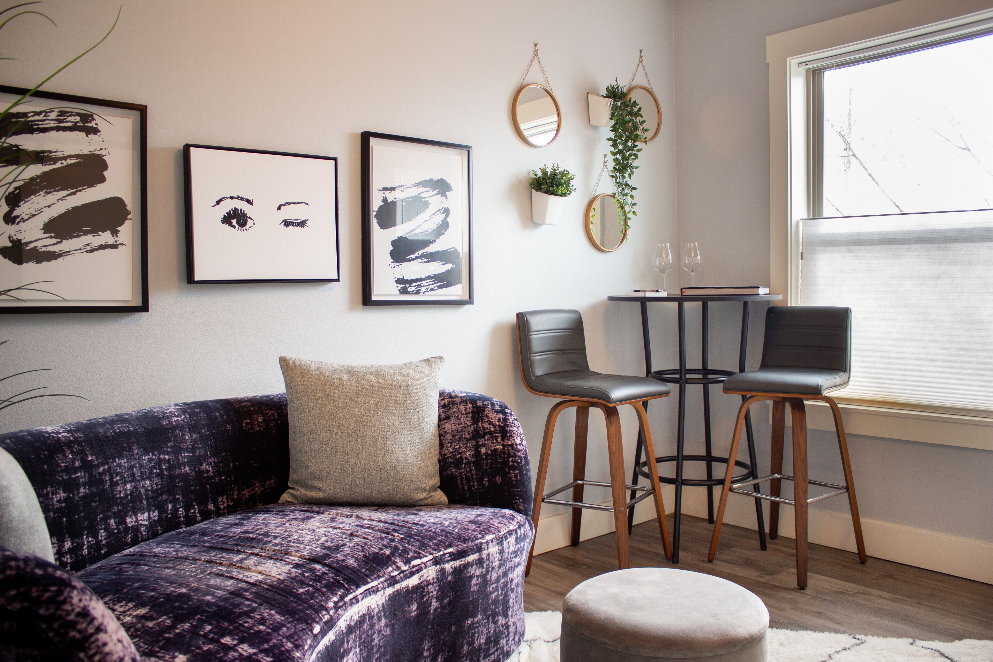 Un salon moderne avec un canapé violet, deux tabourets de bar, une petite table ronde et des œuvres d’art encadrées accrochées au mur.