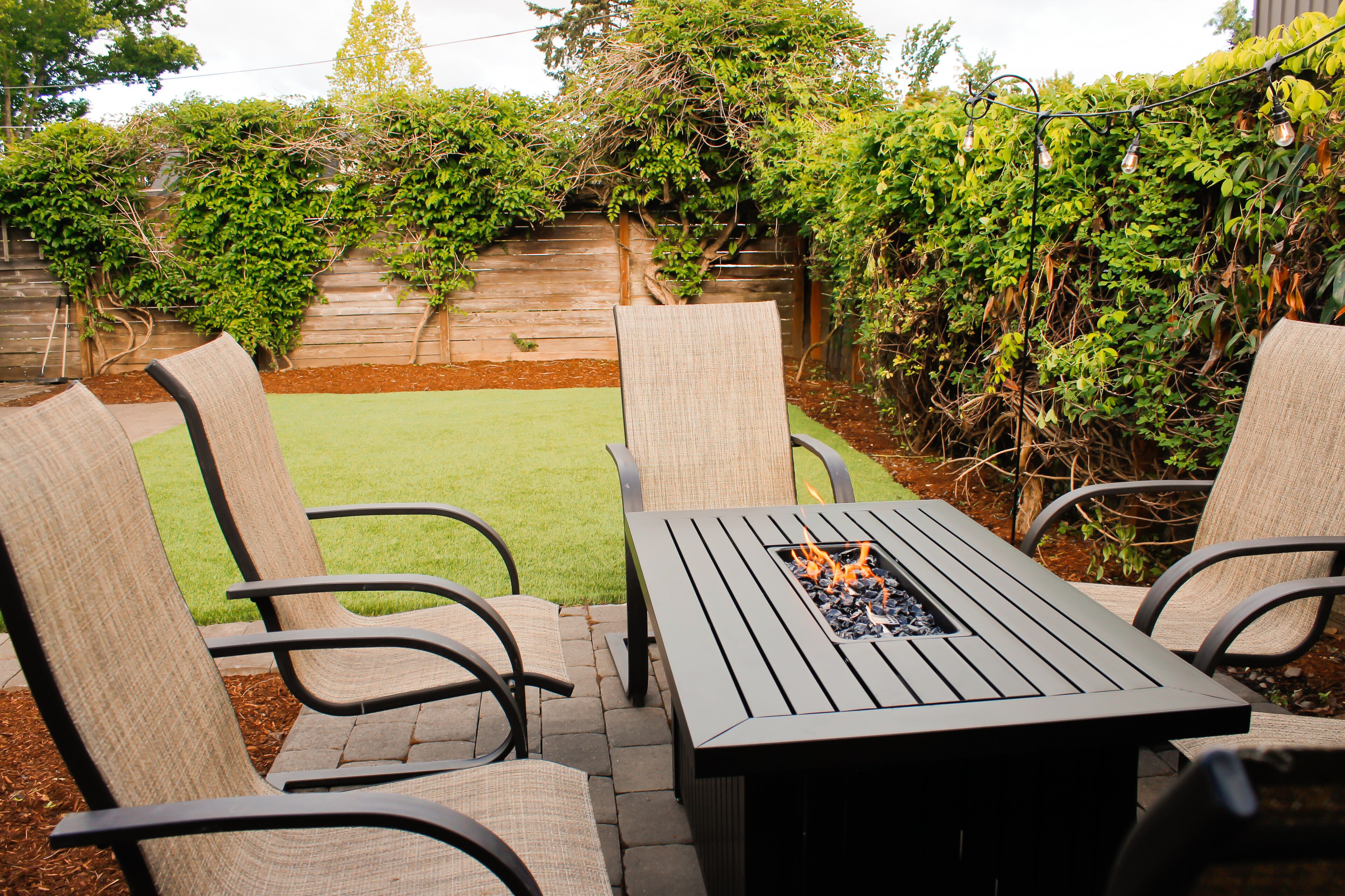 An outdoor patio with a metal table and four chairs, surrounded by a wooden fence and greenery.