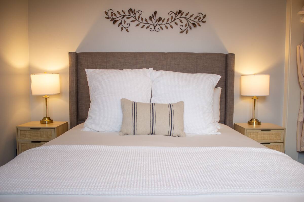 A neatly made bed with white bedding and two pillows, flanked by two bedside tables with lamps, in a room with a decorative wall piece above the bed.