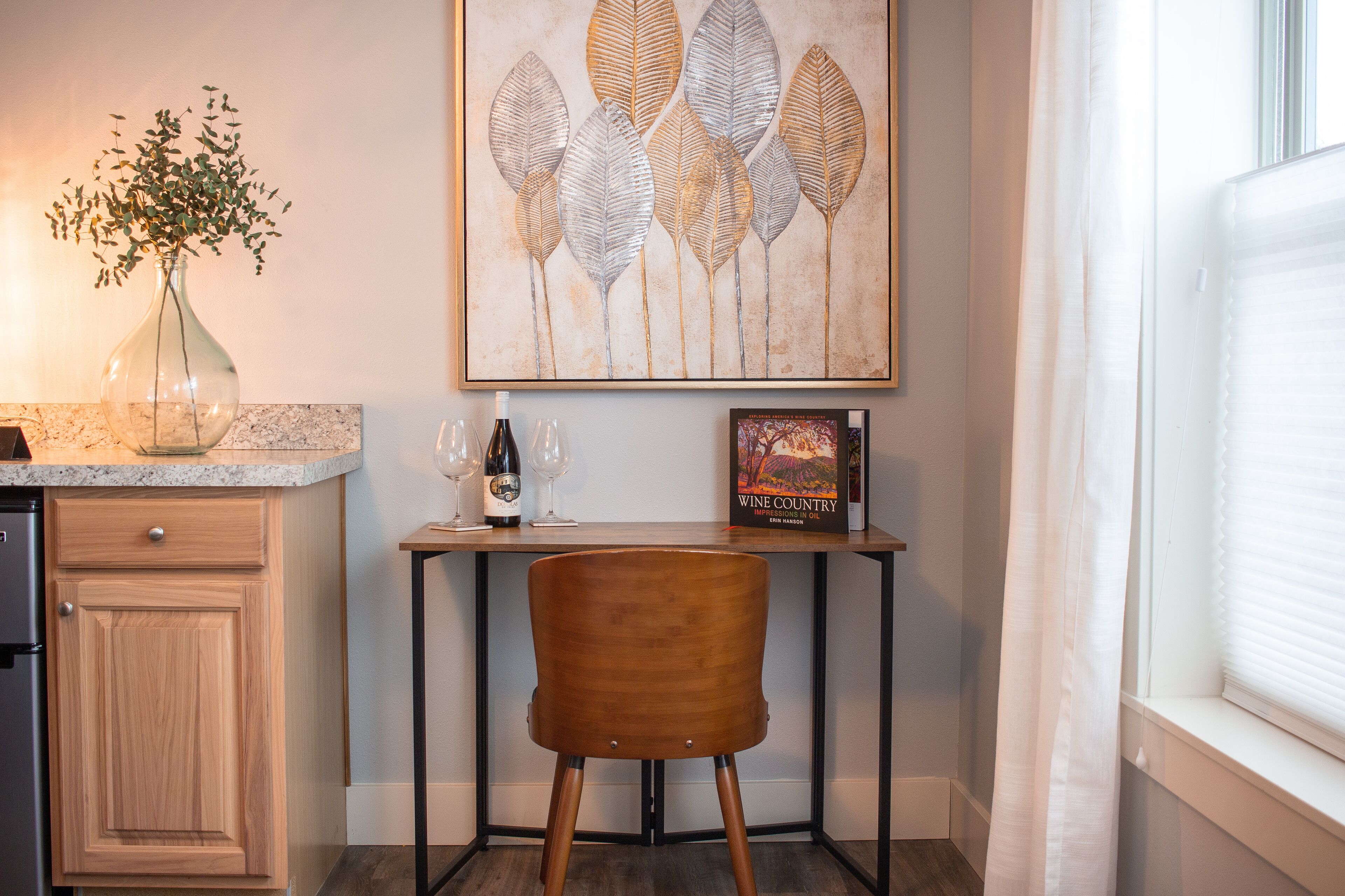 A kitchen with a wooden cabinet, a vase with green leaves, a wine bottle, and a book titled "Wine Country" on a table.