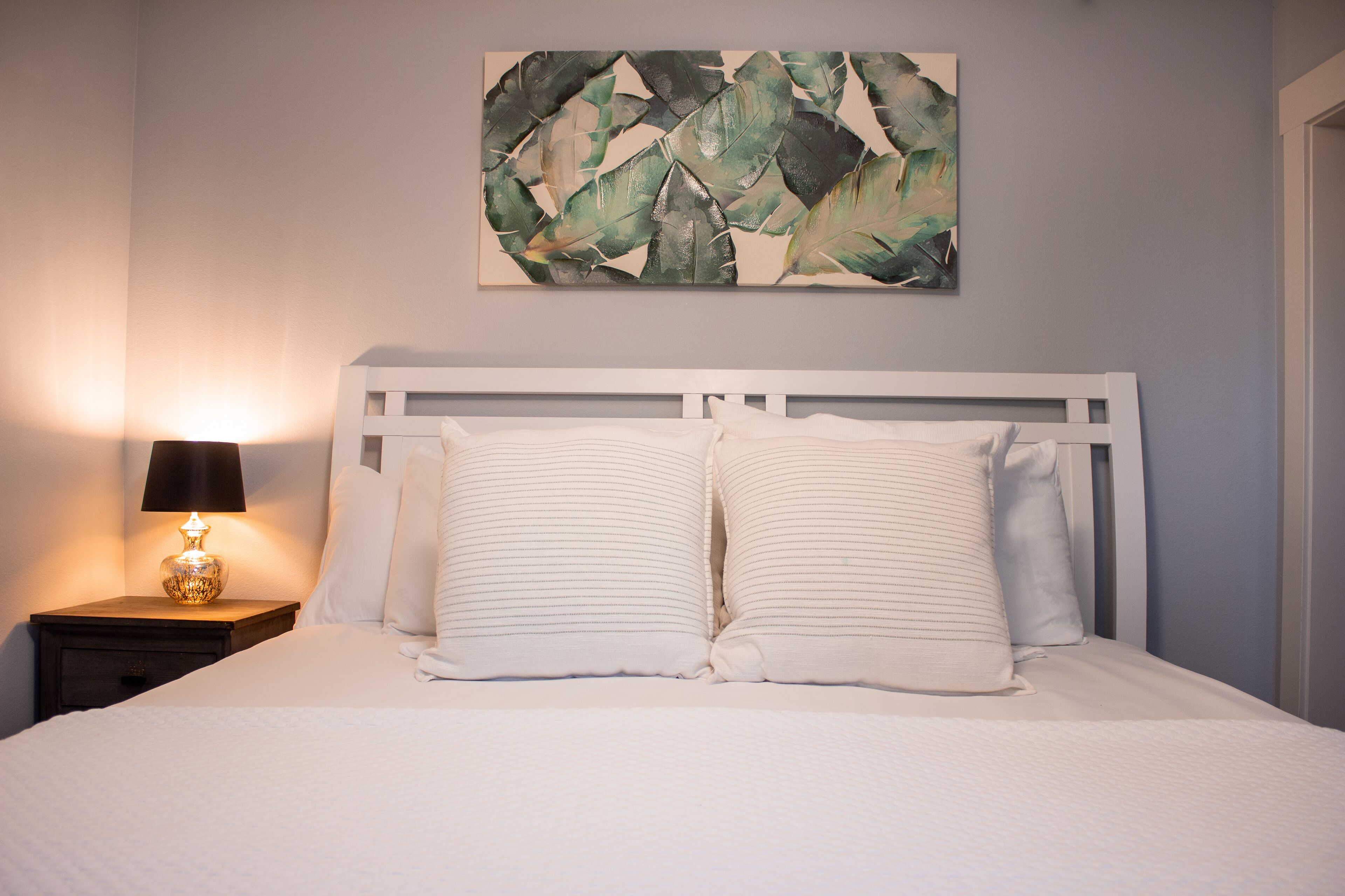 Ein ordentlich bezogenes Bett mit weißen Leinen und gestreiften Kissen, eine Nachttischlampe sowie ein an der Wand hängendes abstraktes Gemälde.