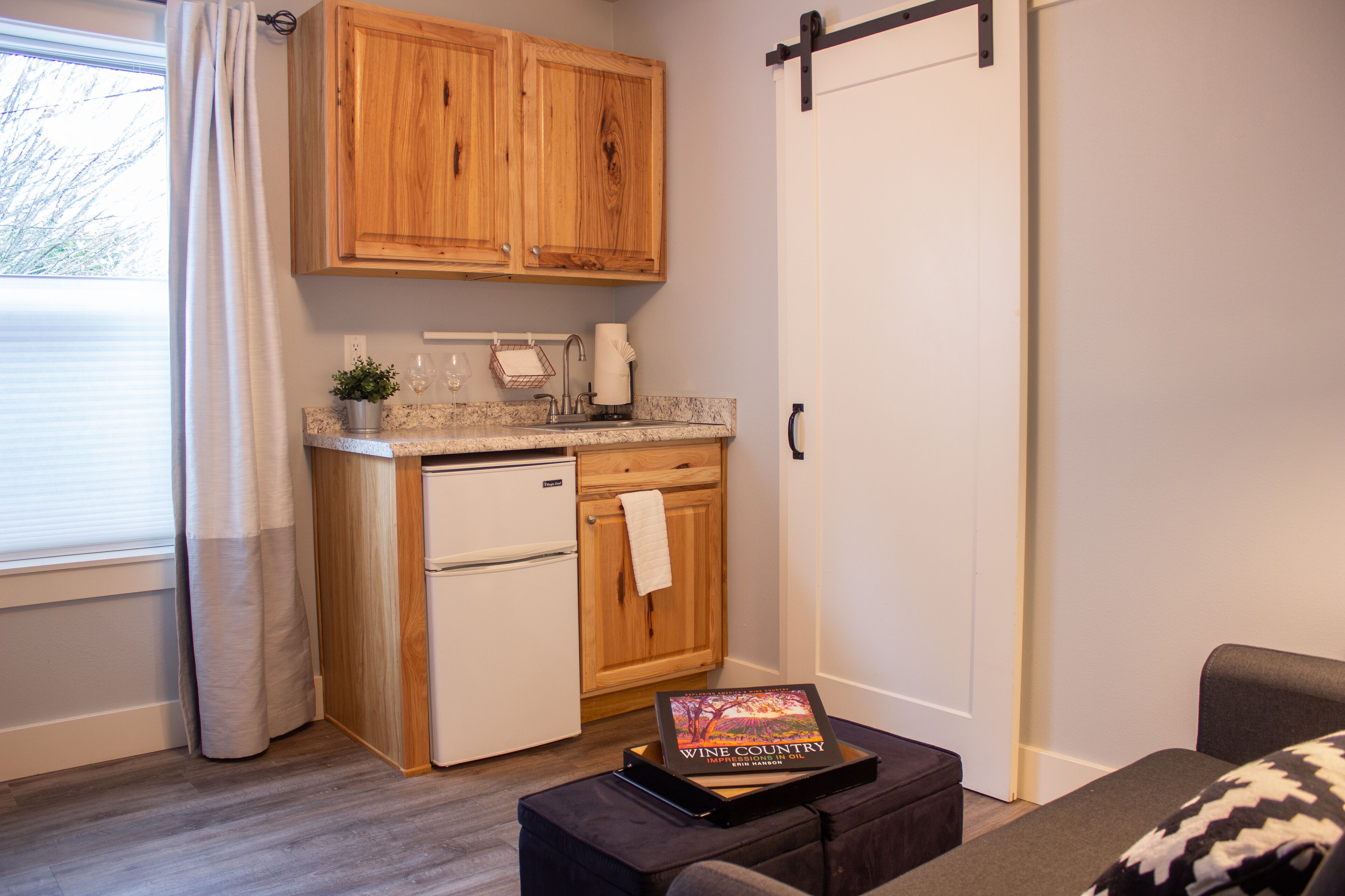 A compact kitchen with wooden cabinets, a small refrigerator, and a countertop with a sink. There's a book titled "Wine Country" on a table, and a sliding door to the right.