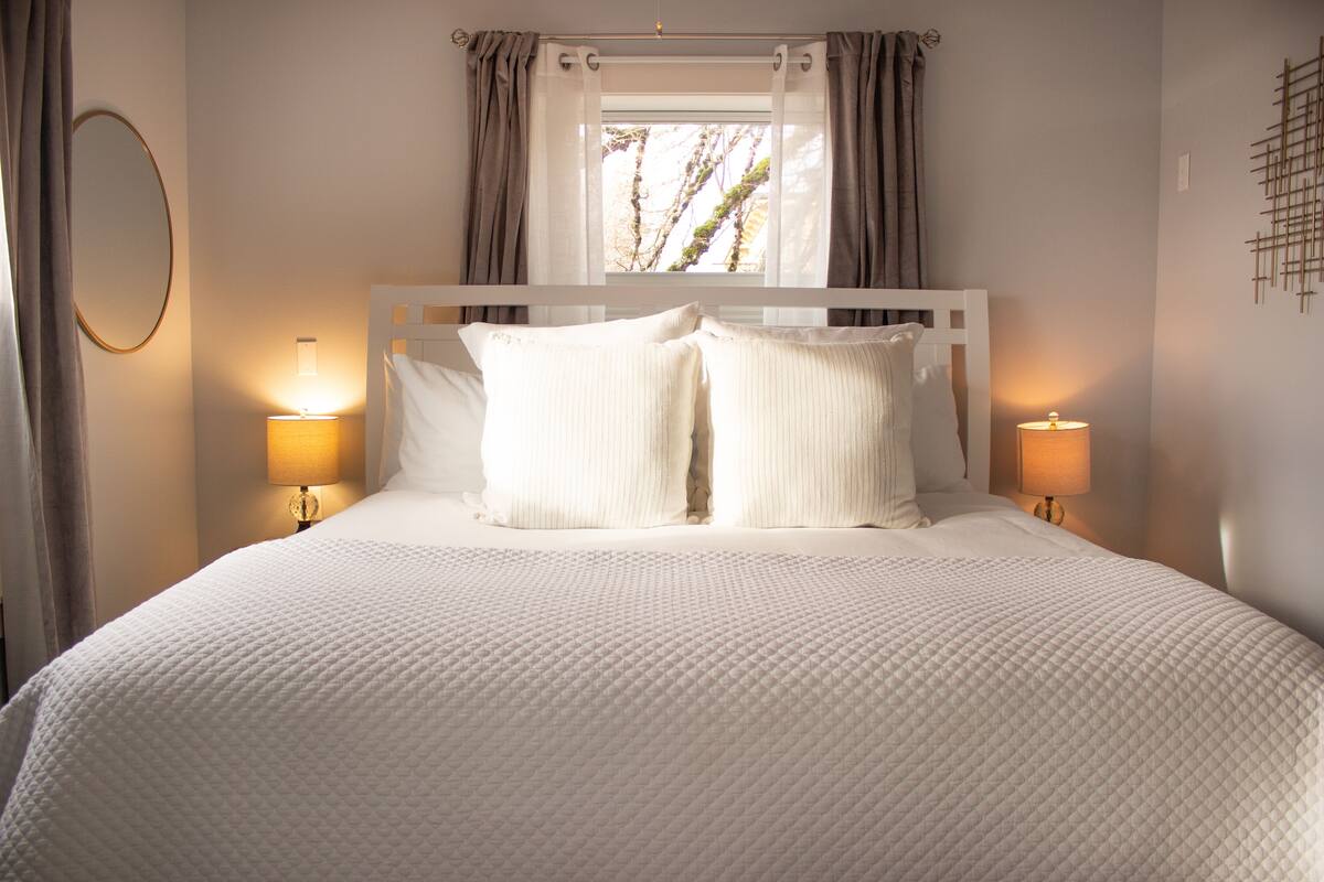 A neatly made bed with a quilted white comforter, two bedside lamps, and a window with curtains.