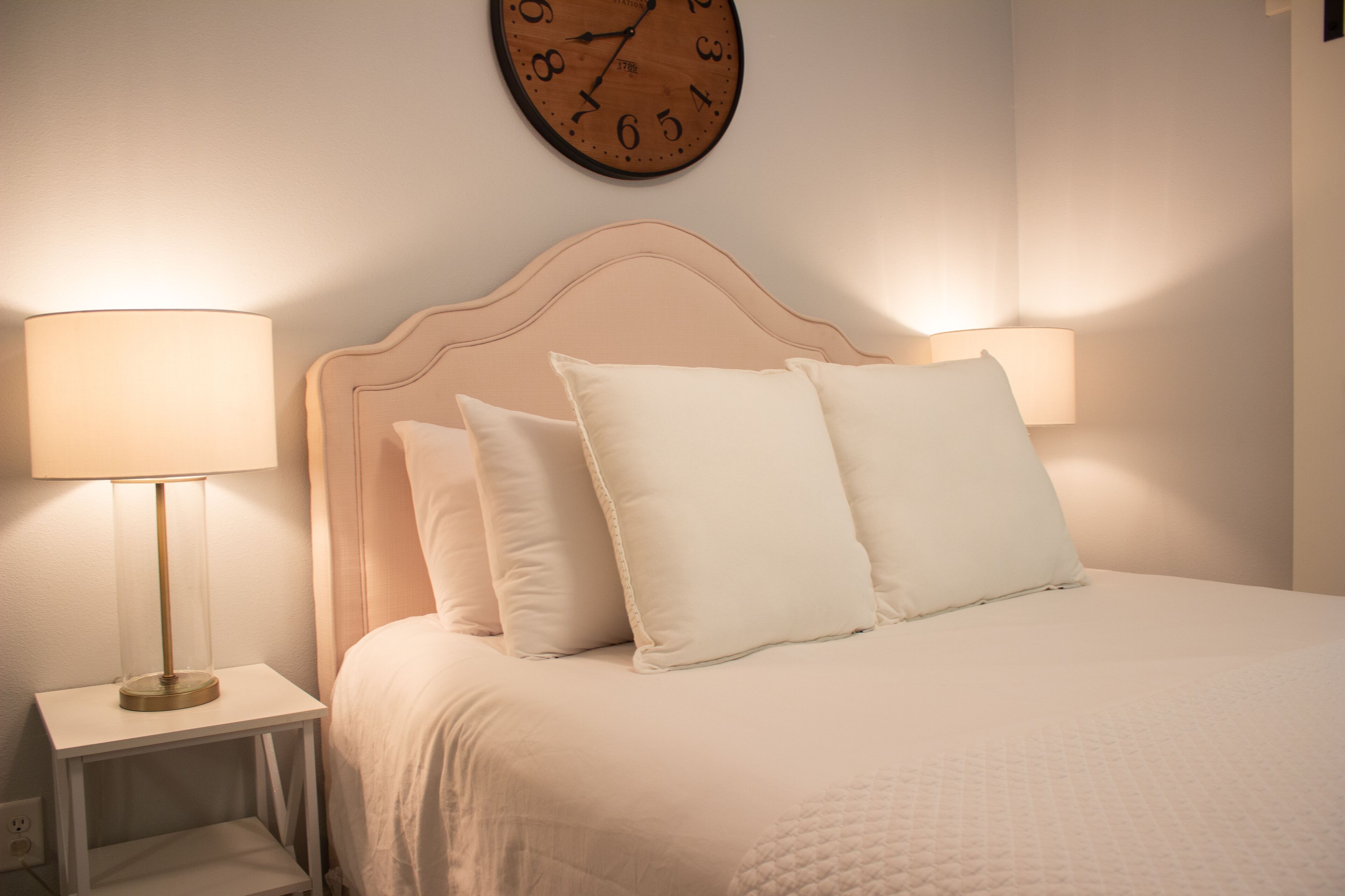 A neatly made bed with a headboard, two bedside lamps, and a wall clock.