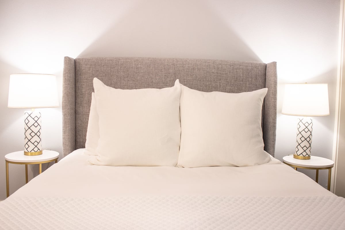 A neatly made bed with a grey headboard, two bedside tables with lamps, and a plain wall.