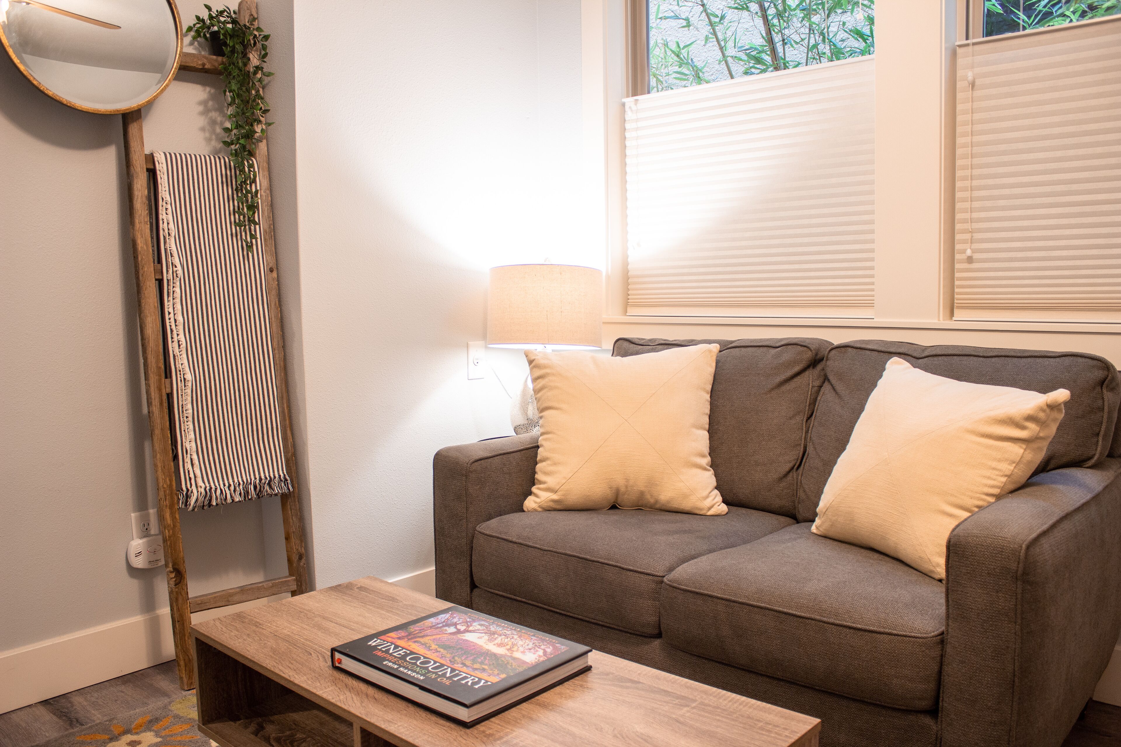 Un salon chaleureux avec un canapé marron, deux coussins, une table basse en bois sur laquelle repose un livre, une lampe et une serviette rayée accrochée à une échelle en bois.