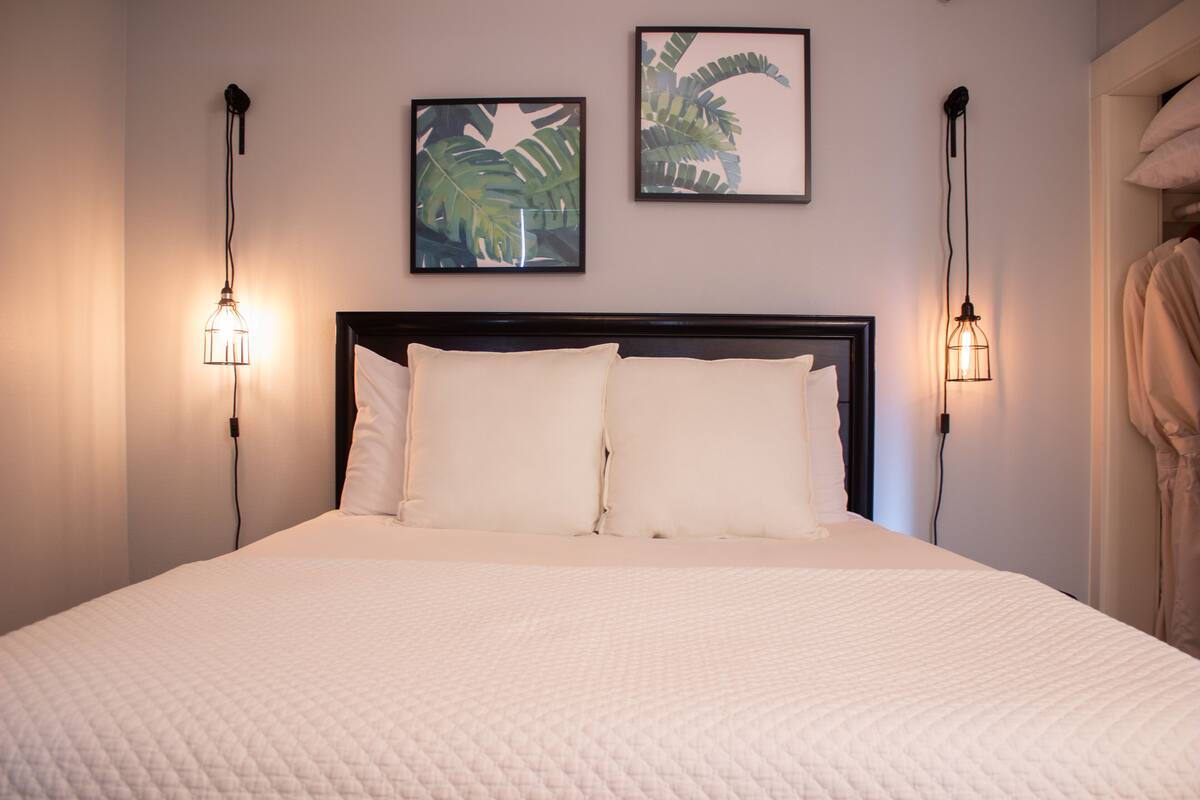 A neatly made bed with a white quilted bedspread, flanked by two wall-mounted light fixtures and framed botanical prints above.