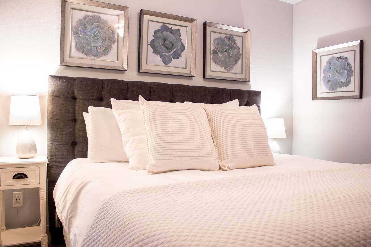 A neatly made bed with a quilted cover, a tufted headboard, and three framed floral artworks above it.