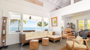 A spacious living room with a large sectional sofa, a wooden coffee table, and a wicker chair. There are large windows offering a view of the ocean and palm trees. - Moorea Beach Lodge (Moorea-Maiao)