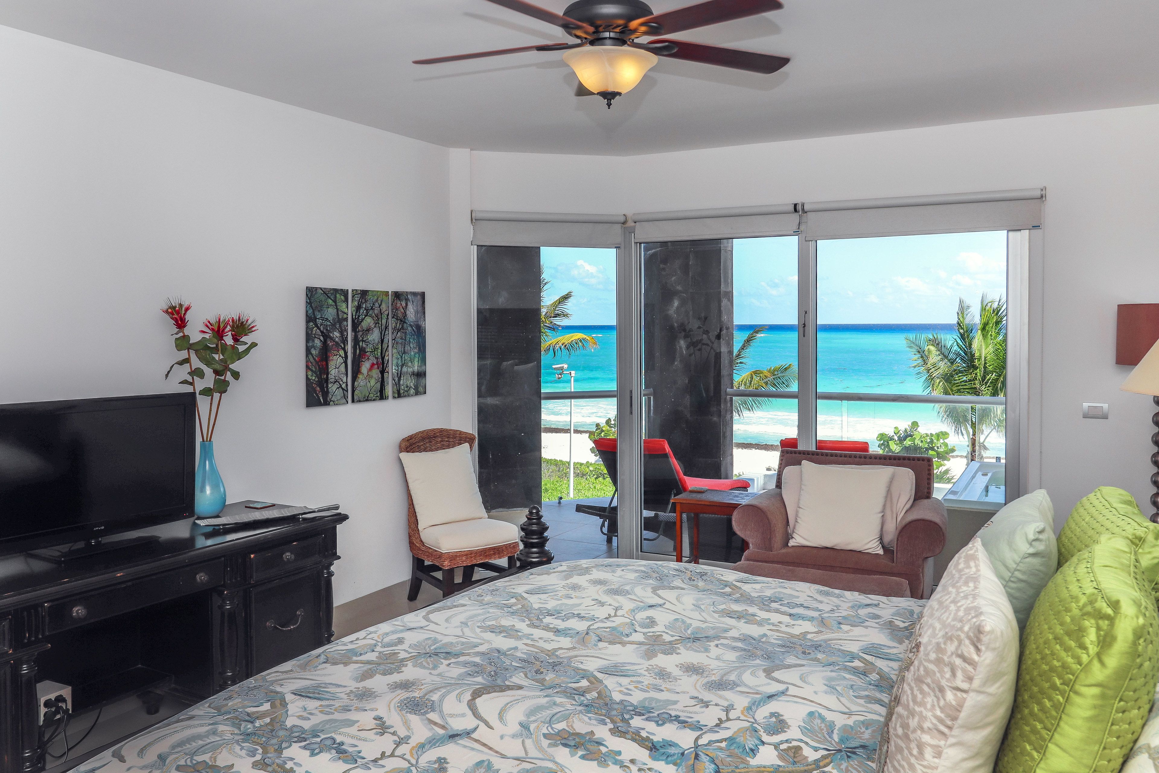 A bedroom with a bed, a TV, a dresser, a chair, a table, and a view of the ocean through large windows.