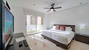 Una habitación de hotel moderna con una cama grande, un escritorio con televisor de pantalla plana, controles remotos, un ventilador de techo y un puerta corrediza de vidrio que da a un balcón.