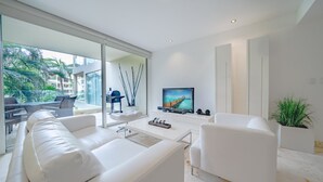 Una sala moderna con un sofá blanco, una mesa de centro y un televisor de pantalla plana sobre un soporte blanco. Hay un ventanal que ofrece vistas al exterior.