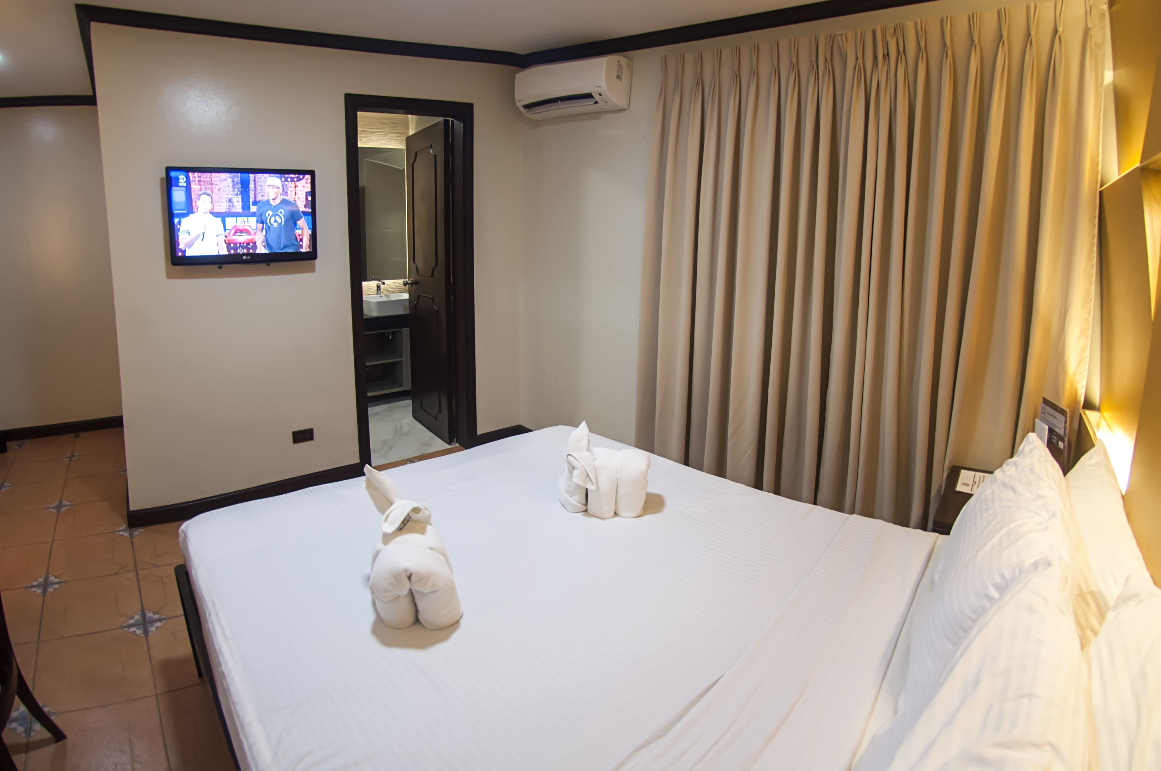 A hotel room with a bed, a television, an air conditioning unit, and a bathroom door.