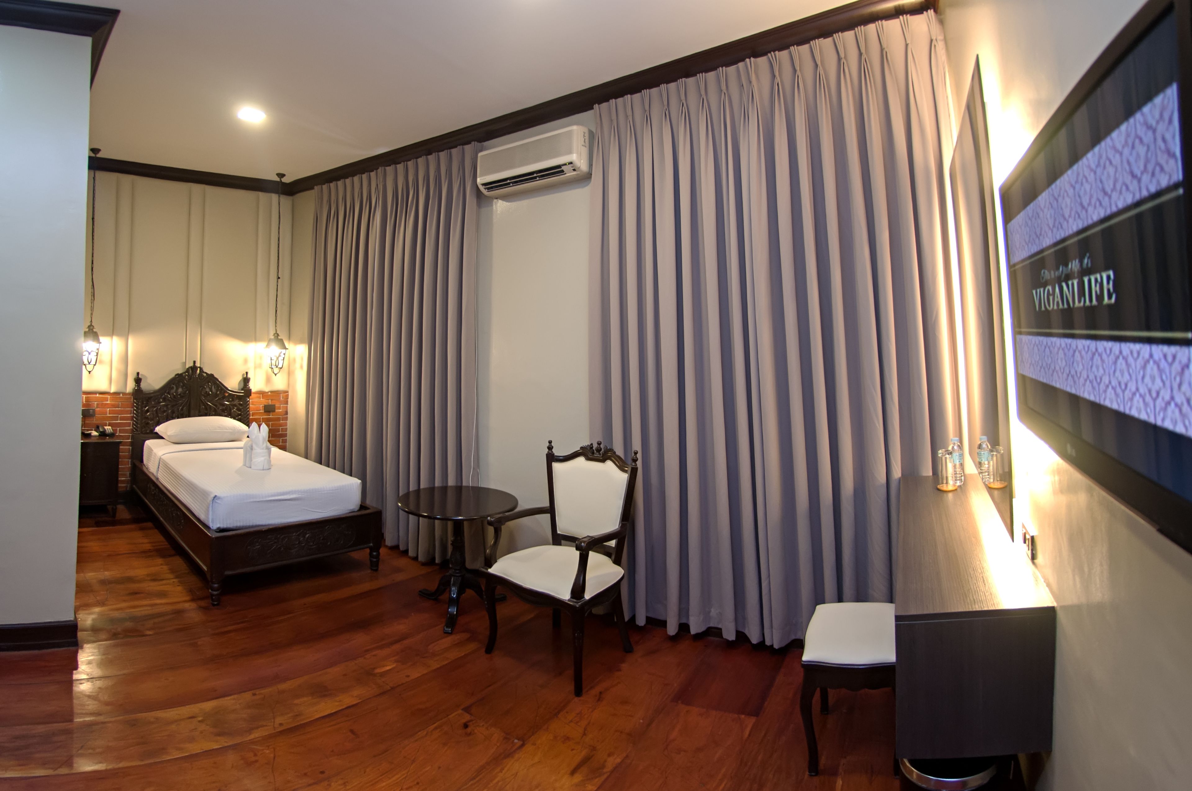A hotel room with a bed, a chair, a small table, an air conditioning unit, and a wall-mounted TV.