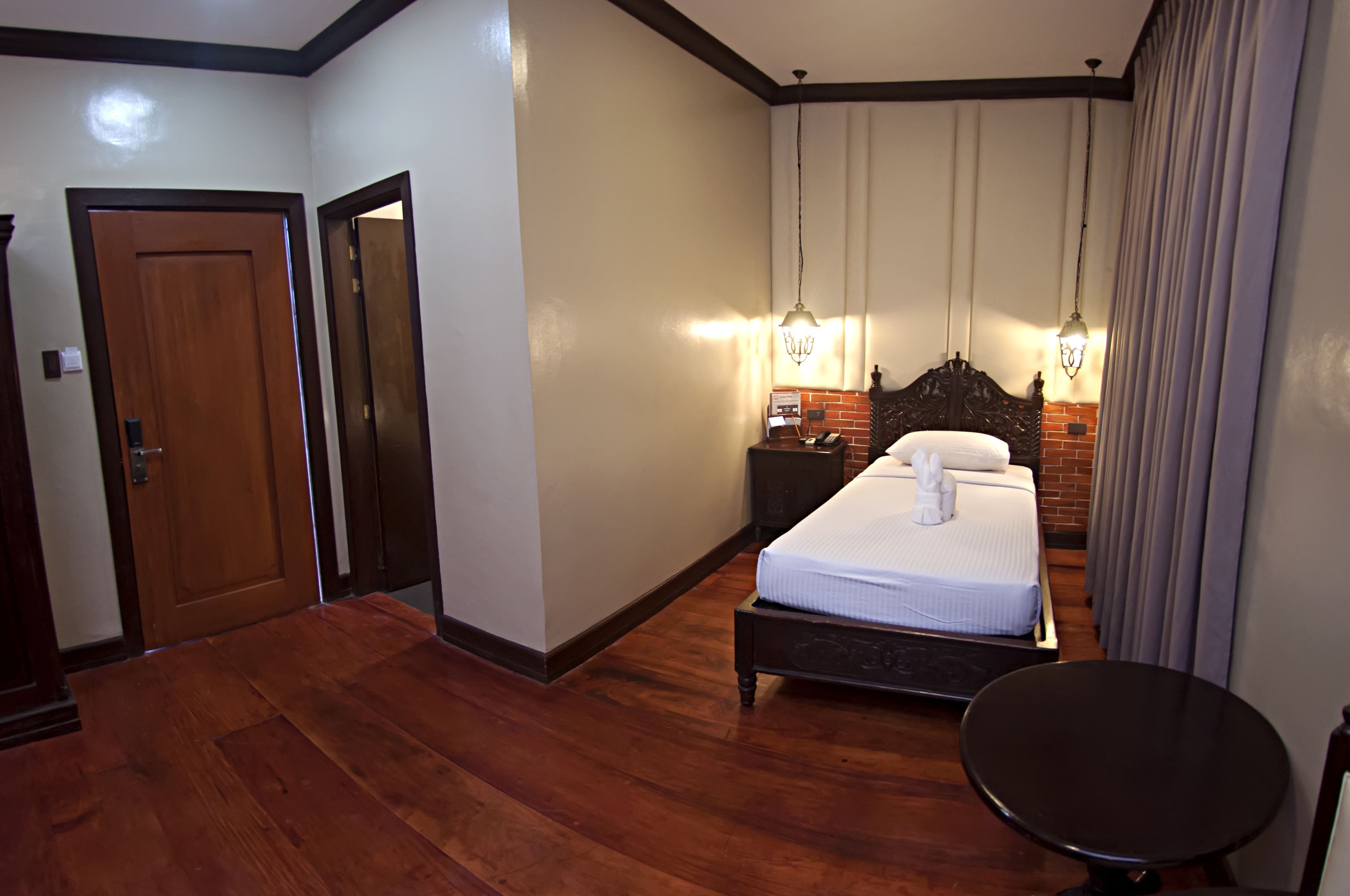 A hotel room with a wooden floor, a bed with white linens, a nightstand, and a hanging light fixture.