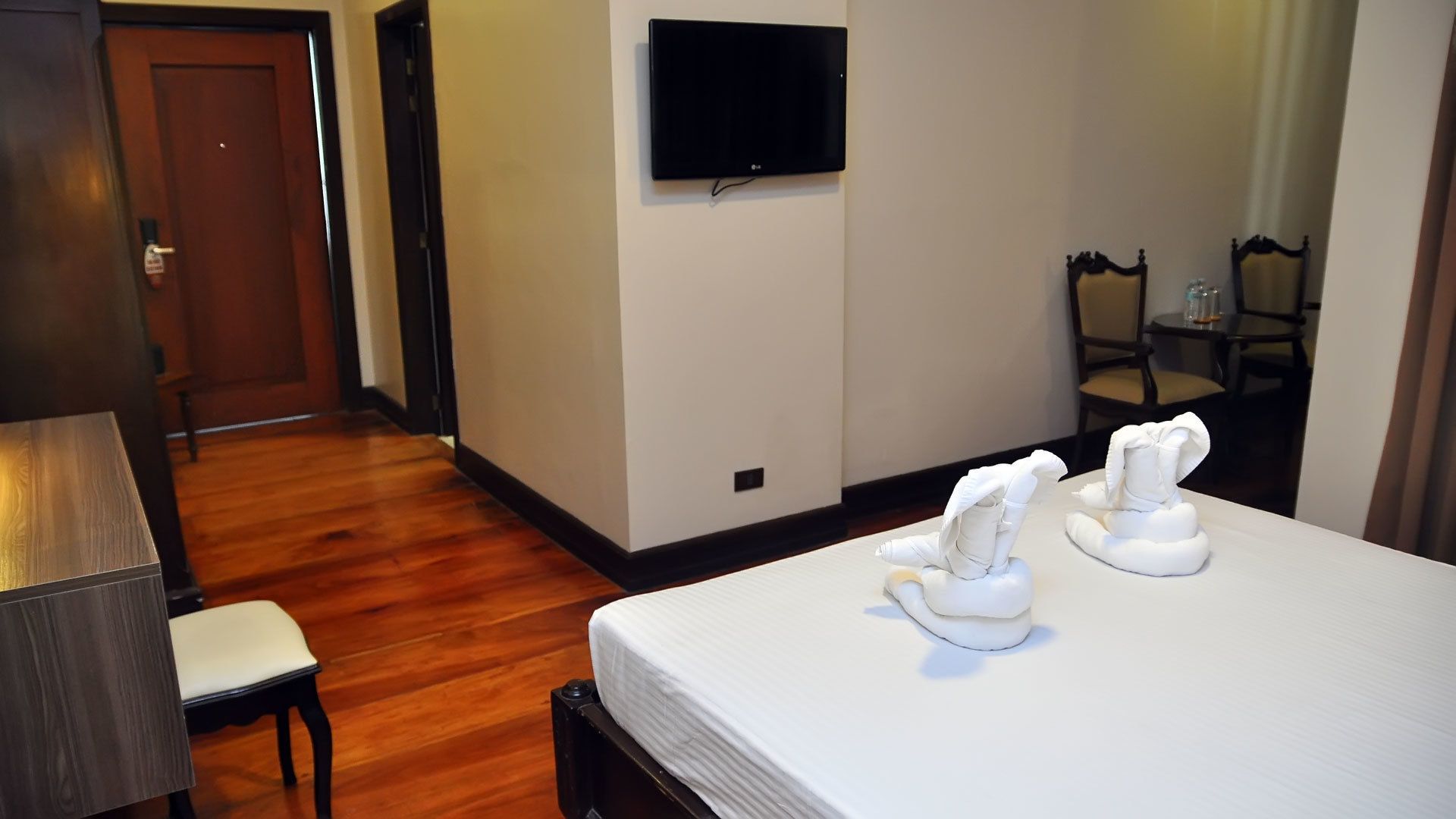 Habitación de hotel con piso de madera, una cama con ropa blanca, un televisor de pantalla plana montado en la pared, una pequeña mesa con sillas y una puerta que da a otra habitación.