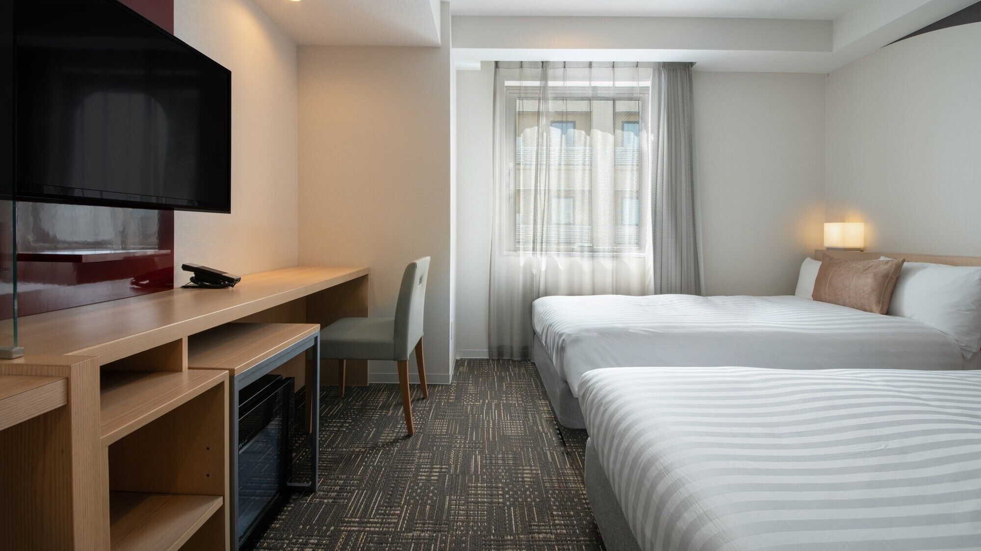 A hotel room with a TV, desk, chair, two beds, and a window with curtains.