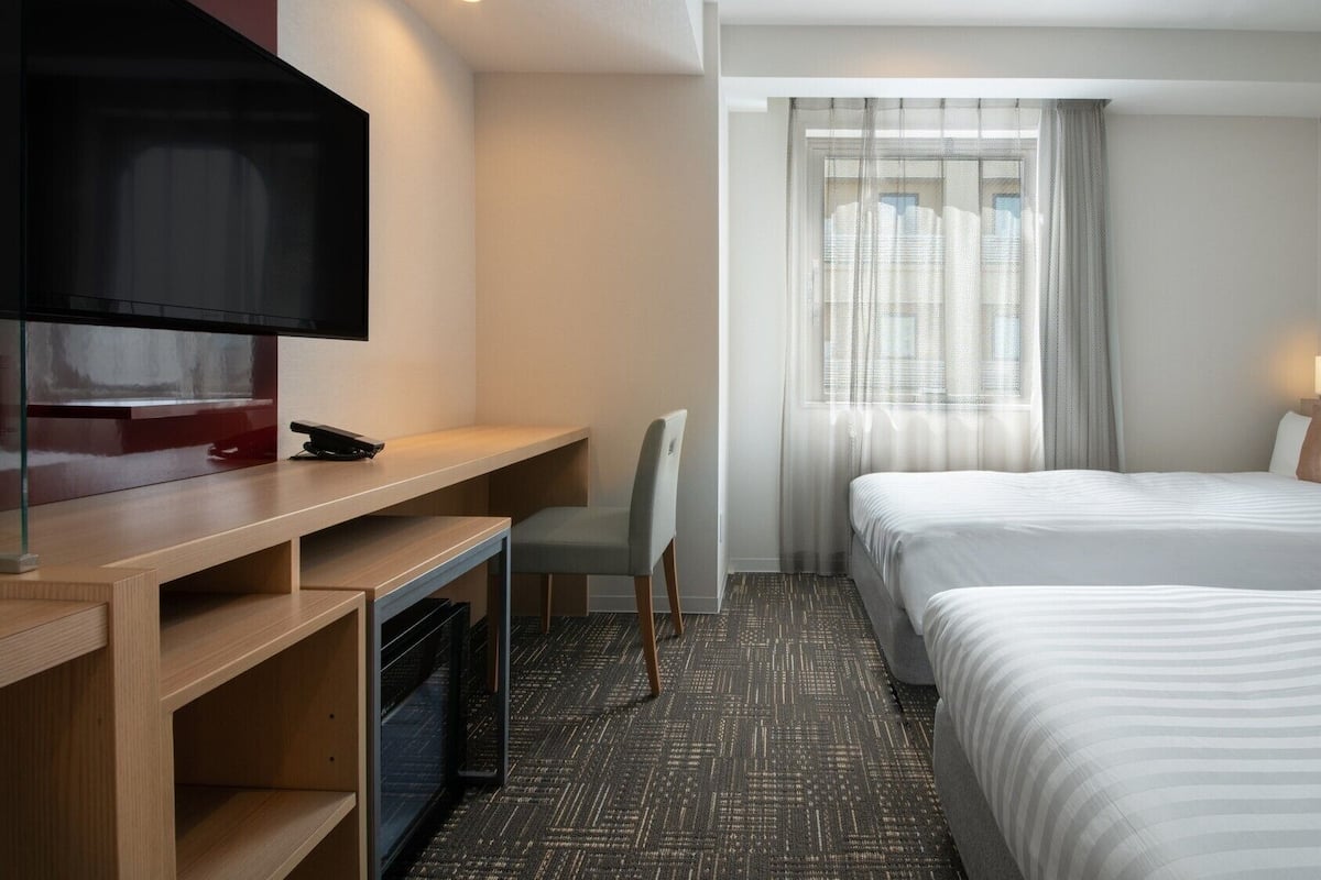 Habitación de hotel con televisión, escritorio, silla, dos camas y ventana con cortinas.