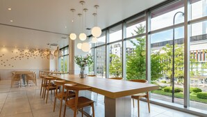 Un comedor moderno con una larga mesa de madera y sillas, ventanas amplias con vistas a zonas verdes y edificios, e iluminación colgante.