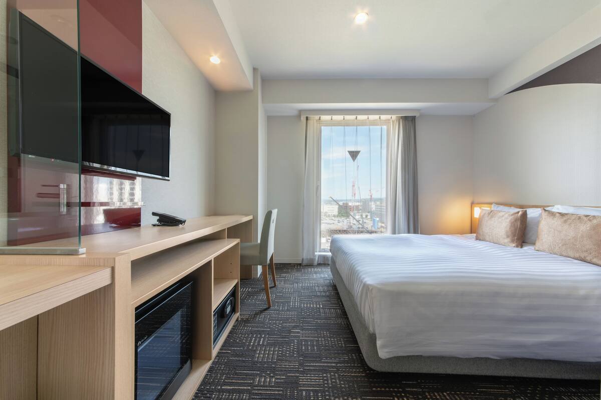Habitación de hotel con cama, escritorio, televisión y vistas a la ciudad desde la ventana.