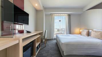 A hotel room with a bed, a desk, a television, and a view of the city through the window.