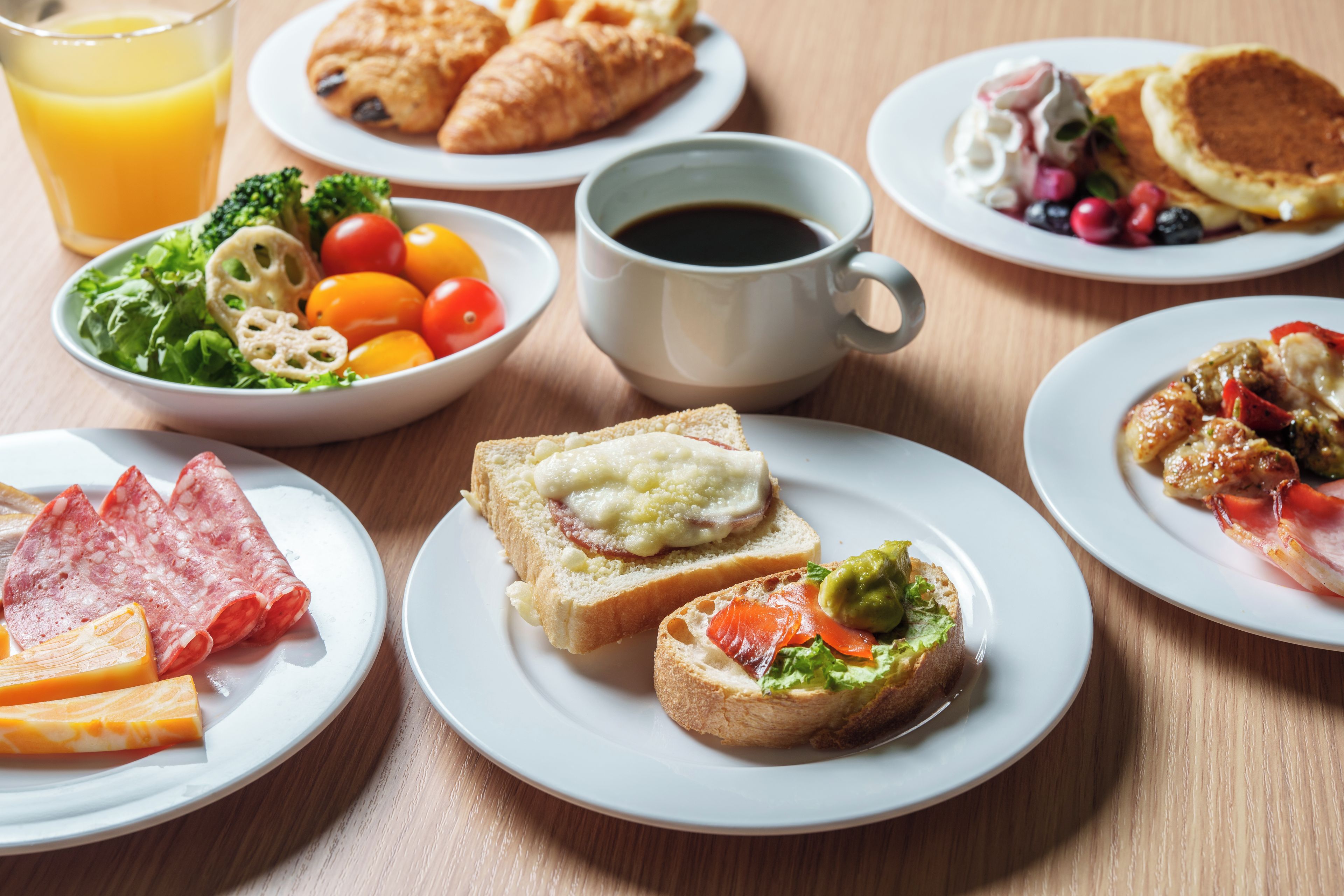 Aamiaispöytä, jossa on monenlaista tarjottavaa, kuten croissantteja, kulho salaattia ja lautasellisia erilaisia ruokia.