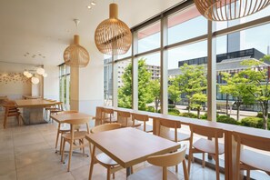 A modern dining area with wooden tables and chairs, large windows offering a view of the cityscape.