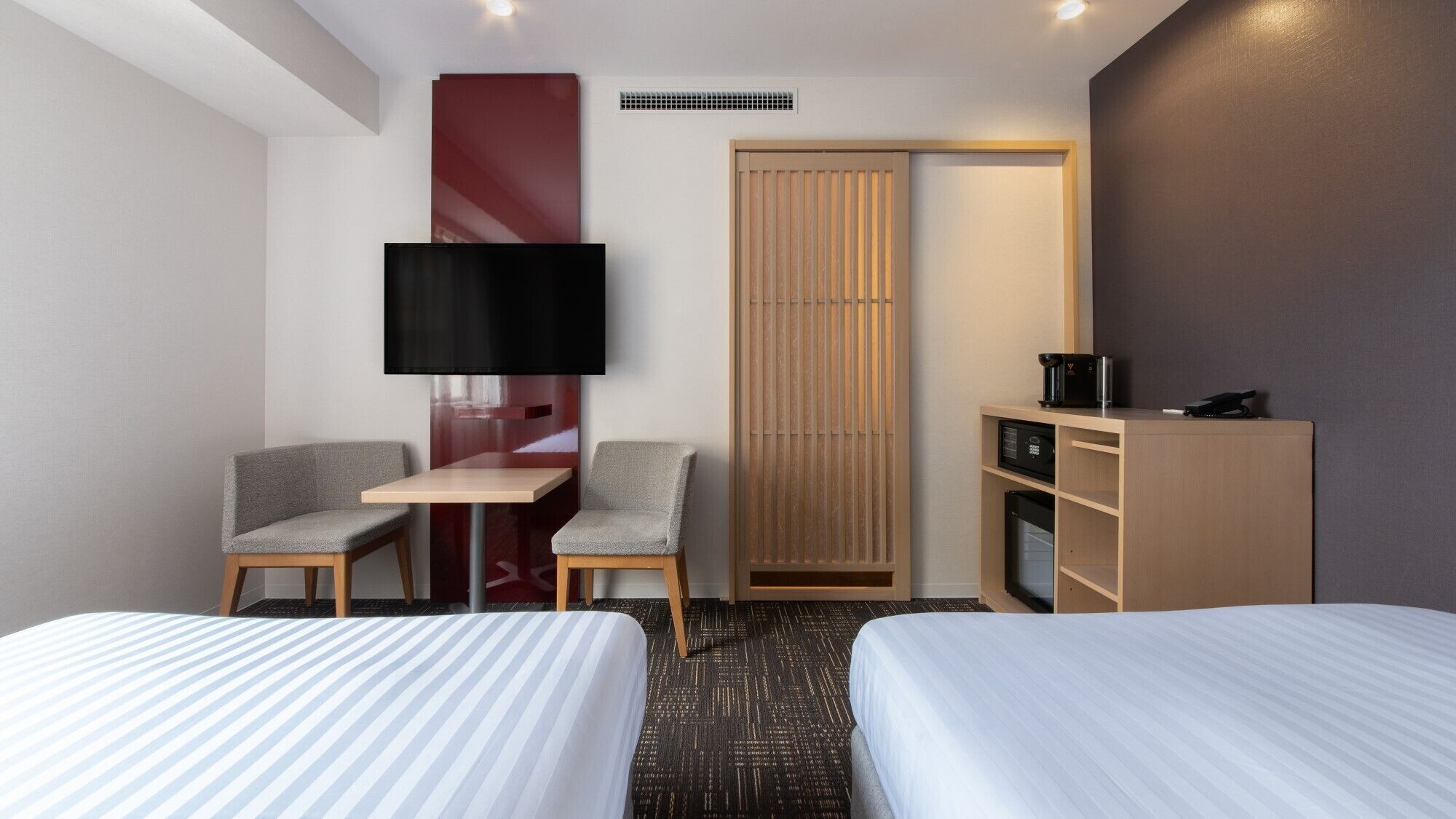 A hotel room with two beds, a desk, a chair, a TV, a minibar, and a sliding door.