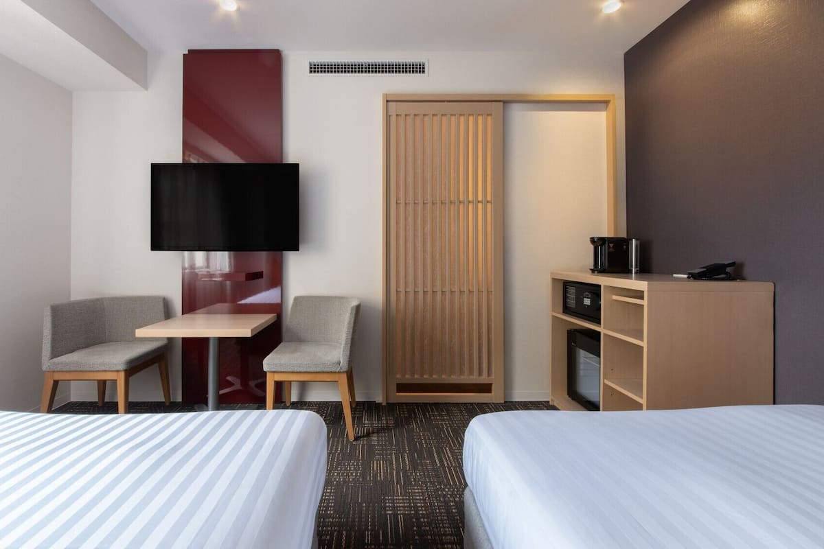 Habitación de hotel con dos camas, un escritorio, una silla, un televisor, un minibar y una puerta corredera.